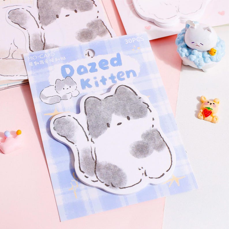 Puede incluir: Un paquete de notas adhesivas "Dazed Kitten" con un dise&ntilde;o de gato de dibujos animados en gris y blanco. El paquete est&aacute; sobre un fondo a cuadros azul, con otros elementos decorativos, incluido un peque&ntilde;o oso con una fresa.