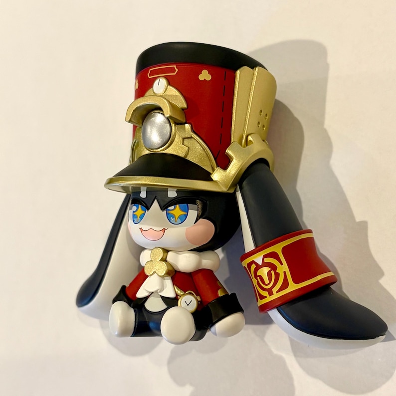 May include: A stylized figurine featuring a character with large star-shaped eyes, wearing a red and gold military-style hat with a silver emblem. The figure has long black ears, a red jacket with white accents, and a small clock detail on the chest.