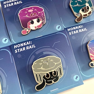 May include: Enamel pins featuring characters from "Honkai: Star Rail." The pins showcase various characters with unique designs and colors, set against a blue background with the game's logo. Each pin is attached to a card.