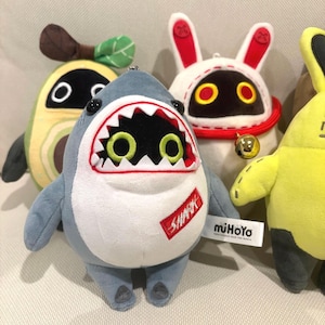 May include: A plush shark toy with a white belly, grey body, and a red tag reading SHARK. It features a wide mouth with sharp teeth and green eyes. Beside it are an avocado plush, a white rabbit-eared plush with a bell, and a yellow plush.