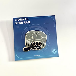 May include: Enamel pin featuring a black cartoon character with large eyes, inside a gray container. The pin is on a blue card with the text "HONKAI: STAR RAIL". The pin has a black outline and is set against a white background.