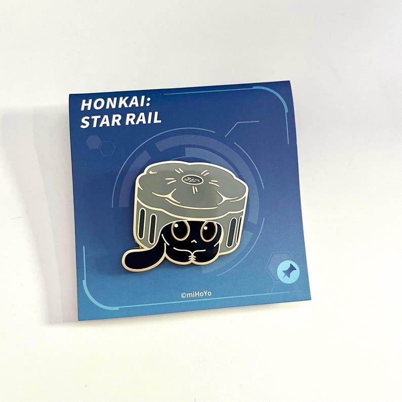 May include: Enamel pin featuring a black cartoon character with large eyes, inside a gray container. The pin is on a blue card with the text "HONKAI: STAR RAIL". The pin has a black outline and is set against a white background.