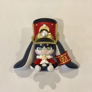 May include: A stylized figurine featuring a character with large blue eyes containing yellow star accents, wearing a red and gold military-style hat, a red cape with a white collar, and long dark blue ear-like appendages with a red cuff on one side.