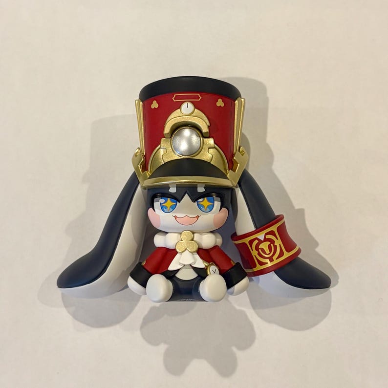 May include: A stylized figurine featuring a character with large blue eyes containing yellow star accents, wearing a red and gold military-style hat, a red cape with a white collar, and long dark blue ear-like appendages with a red cuff on one side.