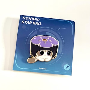 May include: A pin featuring a cartoon cat wearing a sushi-shaped hat. The pin is on a blue card with the text "HONKAI: STAR RAIL". The cat has large eyes and a striped tail. The sushi hat is purple with a gold center.