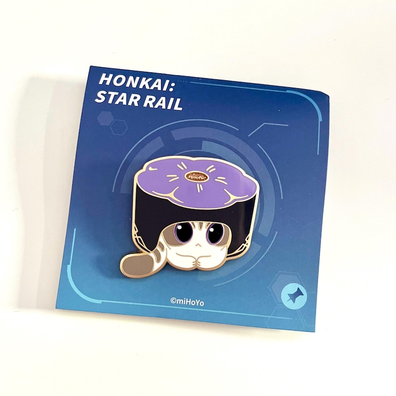 May include: A pin featuring a cartoon cat wearing a sushi-shaped hat. The pin is on a blue card with the text "HONKAI: STAR RAIL". The cat has large eyes and a striped tail. The sushi hat is purple with a gold center.