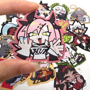 Könnte beinhalten: Eine Sammlung farbenfroher Aufkleber im Anime-Stil. Der zentrale Aufkleber zeigt einen pinkhaarigen Charakter mit einem verspielten Ausdruck, umgeben von anderen Charakterdesigns. Die Aufkleber werden von einer Hand gehalten.