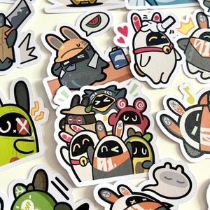 May include: A collection of colorful stickers featuring cartoon rabbit characters in various poses and outfits. Some stickers include speech bubbles, musical notes, and other graphic elements. The stickers have a white border and are arranged on a light-colored surface.