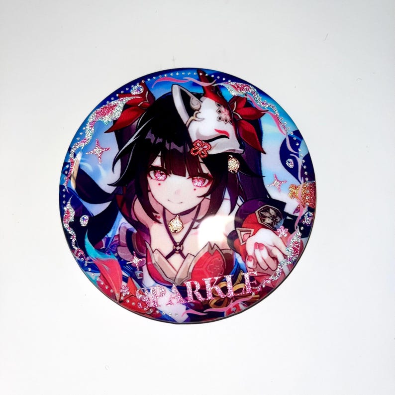 May include: A round button with a colorful anime character with long black hair and a red and white mask. The character is wearing a red and gold outfit and has pink glitter around her. The button has a pink glitter border and the word "SPARKLE" written in pink glitter.