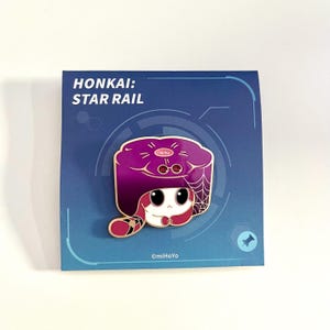 May include: A purple and white enamel pin featuring a cartoon character from Honkai: Star Rail. The character has a purple hat with a spiderweb design and a white face. The pin is on a blue card with the text "HONKAI: STAR RAIL".