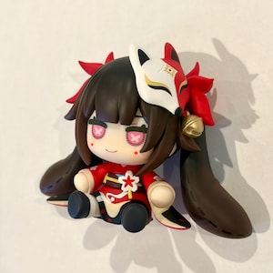 May include: A chibi-style figurine featuring a character with dark brown hair, wearing a red and black outfit with a white flower accessory. The figure has a white and red fox mask perched on its head, pink cross-shaped eyes, and a small gold bell.