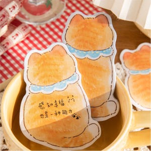 Puede incluir: Notas adhesivas con forma de gato naranja con collares azules y texto en chino. Las notas est&aacute;n dentro de una taza amarilla. Las notas tienen un efecto acuarela y huellas de patas.