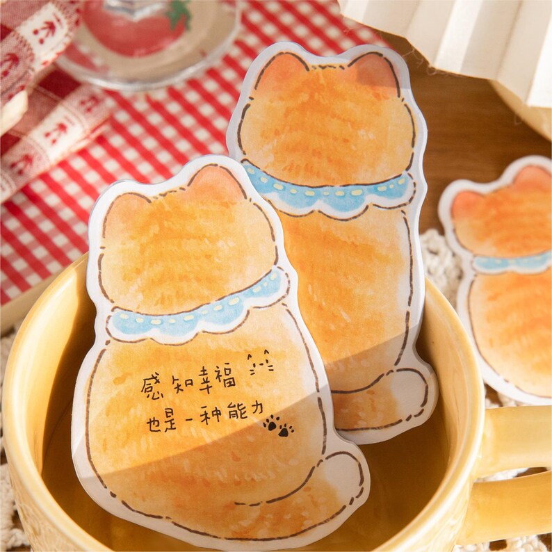 Puede incluir: Notas adhesivas con forma de gato naranja con collares azules y texto en chino. Las notas est&aacute;n dentro de una taza amarilla. Las notas tienen un efecto acuarela y huellas de patas.