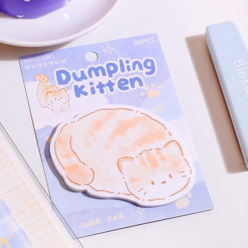 Puede incluir: Un paquete de notas adhesivas "Dumpling Kitten". Las notas tienen forma de gato y tienen rayas naranjas y blancas. El paquete es azul claro con el texto "Dumpling Kitten" en azul y amarillo. El paquete tambi&eacute;n tiene el texto "30PCS".