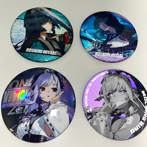 Puede incluir: Cuatro botones redondos de estilo anime con efectos holográficos. Dos botones presentan a una mujer con pelo largo negro y el texto "HOSHIMI MIYABI". Los otros dos muestran a una mujer con pelo morado y el texto "MOCKING BIRD".