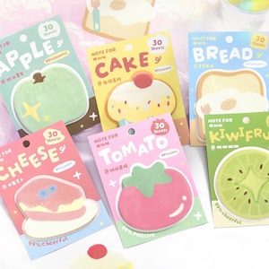 Puede incluir: Un conjunto de blocs de notas adhesivas de colores con forma de alimentos, como una manzana, un pastel, pan, queso, tomate y kiwi. Cada bloc tiene 30 hojas y presenta textos e ilustraciones.