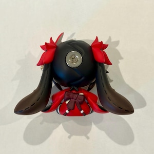 May include: A small figurine featuring a black head with red hair accents and long, dark brown ears. The figure wears a red garment with a dark brown bow on the back. A circular metallic screw is visible on the top of the black head piece.