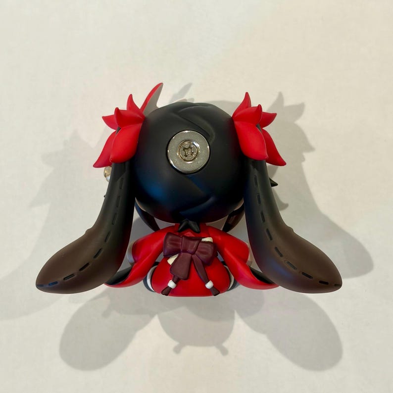 May include: A small figurine featuring a black head with red hair accents and long, dark brown ears. The figure wears a red garment with a dark brown bow on the back. A circular metallic screw is visible on the top of the black head piece.