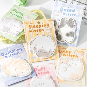 Könnte beinhalten: Eine Auswahl an Haftnotizen in Katzenform mit verschiedenen Designs und Farben. Die Notizen zeigen Illustrationen von Katzen und Sätze wie "Plush Kitten" und "Sleeping Kitten". Jede Packung enthält 30 Haftnotizen.