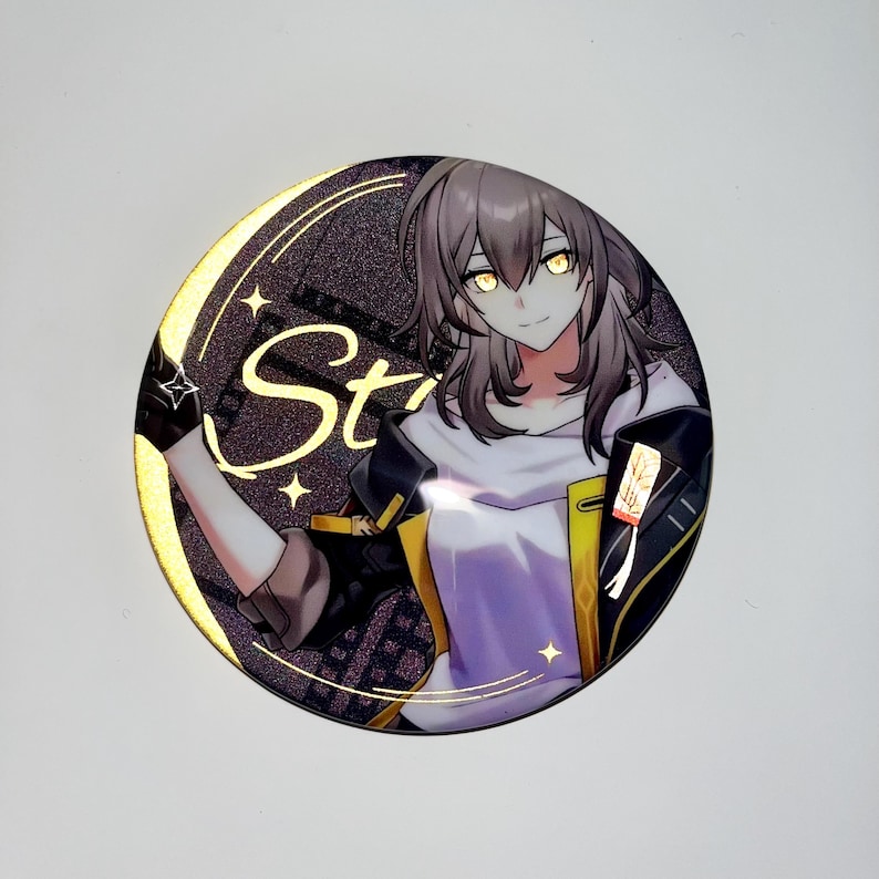 May include: A round button featuring a cartoon anime character with brown hair, yellow eyes, and a white shirt. The character is wearing a black jacket with gold accents and a yellow sash. The background is a gold glitter design with the word "Star" written in gold.