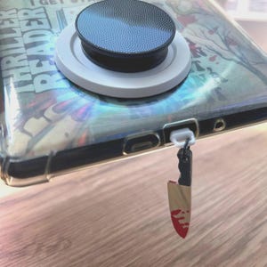 May include: A clear phone case with a pop socket and a small knife charm. The pop socket is black and white. The knife charm has a gold blade with red blood splatter. The phone case has the words "Thriller Reader" on it.