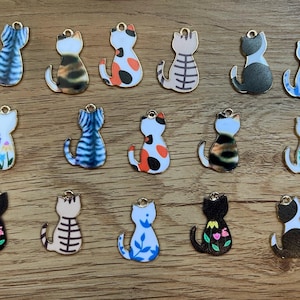 May include: A collection of cat-shaped charms in various colors and patterns. Some charms feature stripes, spots, or floral designs. Each charm has a gold-colored loop at the top for attaching to jewelry or other crafts. The charms are arranged on a wooden surface.