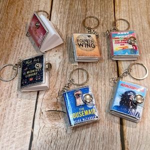 May include: Miniature book keychains with silver key rings. The book covers feature titles like "Tenant", "Fourth Wing", "The Midnight Library", "The Housemaid", "Say You Remember Me", and "Atmosphere". The keychains are on a wooden surface.