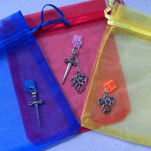 May include: Three small, colorful mesh bags in blue, red, and yellow. Each bag holds a silver-toned earring with a dragon charm and a sword charm. The earrings have a clip-on style.