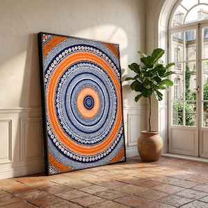 May include: A framed abstract art print with a circular design in orange, navy blue, and white. The artwork is leaning against a wall in a room with a plant in a terracotta pot. The room has a tiled floor.