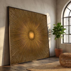 May include: Large, framed artwork featuring a sunburst design. The artwork has a central circular element with radiating lines in shades of gold and brown. The frame is dark, and the piece leans against a wall in a room with natural light.