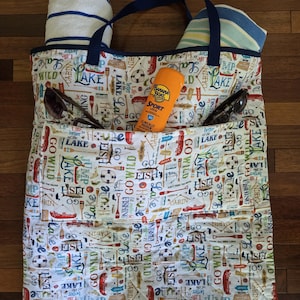 May include: A cream-colored tote bag with a repeating pattern of lake-themed icons and text including "Lake," "Go Wild," and "Fish." The bag features dark blue straps, two rolled towels, a pair of sunglasses, and a bottle of Banana Boat Sport sunscreen.
