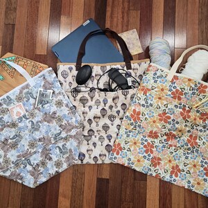 May include: Three fabric tote bags with white or brown handles. The left bag has a blue and brown paisley print, the center bag features a hot air balloon pattern, and the right bag displays a colorful floral print. Each bag has exterior pockets.