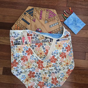 May include: A floral print tote bag with cream-colored straps, holding a wooden board game, playing cards, and a small blue pouch with golf tees. The bag's pattern features orange, blue, and yellow flowers on a white background.