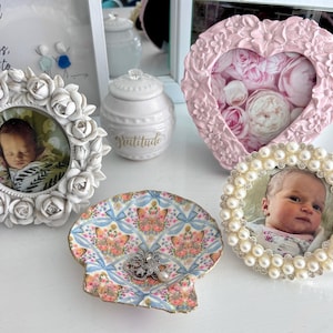 May include: A selection of decorative items, including framed photographs, a seashell-shaped dish, and a small jar. The frames are decorated with roses and pearls. The seashell dish has a floral pattern and holds a piece of jewellery.