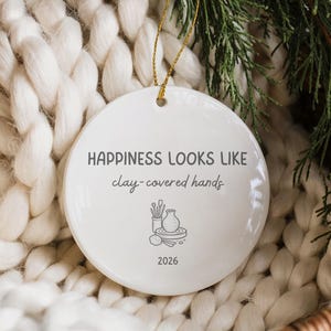 Könnte beinhalten: Ein rundes, weißes Keramikornament mit dem schwarzen Text "HAPPINESS LOOKS LIKE clay-covered hands" über einer Strichzeichnung von Töpferwerkzeugen und einer Vase, mit der Jahreszahl "2026" unten und einer goldenen Aufhängeschnur.