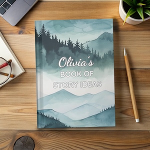 May include: A journal titled "Olivia's Book of Story Ideas" with a watercolor mountain and forest design in shades of teal and gray. The notebook is on a wooden surface, with a pen, glasses, a laptop, and a small plant nearby.