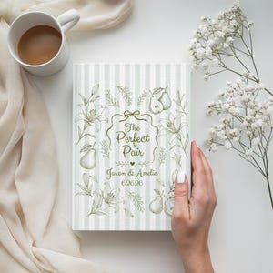 May include: A book with a light green and white striped cover, featuring the text "The Perfect Pair" and the names "Jason & Amelia" with the date "6.26.26". The cover is decorated with floral and pear illustrations.