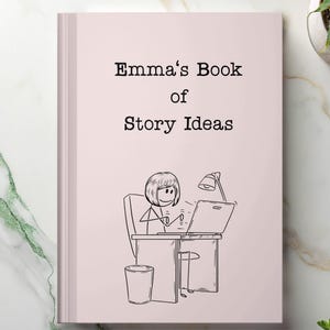 May include: A light pink book titled "Emma's Book of Story Ideas" with a black and white illustration of a person working on a laptop. The illustration shows a desk, chair, and trash can. A small plant is visible in the background.