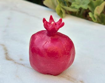 May include: A vibrant red pomegranate-shaped candle. The candle has a textured surface and a detailed crown. The candle is set against a white marble surface with green foliage in the background. The candle is a decorative item.