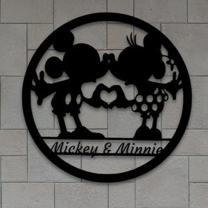 Könnte beinhalten: Schwarze Metall-Wandkunst mit Mickey und Minnie Mouse, die Hände in Herzform halten. Das Design befindet sich in einem kreisförmigen Rahmen, darunter steht "Mickey & Minnie". Die Kunst ist an einer hellgrauen Backsteinmauer befestigt.