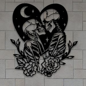 Könnte beinhalten: Schwarze Metall-Wandkunst mit einem herzförmigen Design, das zwei sich umarmende Skelette zeigt. Das Design umfasst einen Mond, Sterne und florale Elemente. Montiert an einer hellgrauen Backsteinmauer, mit grünem Laub am unteren Rand.