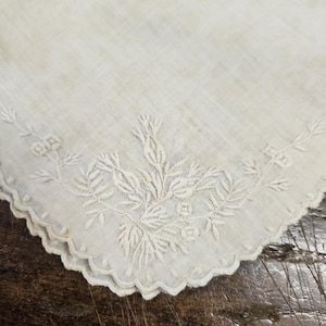 Puede incluir: Pañuelo blanco roto con diseño floral y de hojas bordado. Los bordes son festoneados. El bordado está detallado con una variedad de puntadas, creando una apariencia texturizada. El pañuelo probablemente está hecho de una tela suave y ligera.