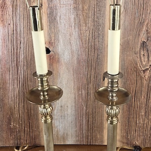 May include: Pair of vintage brass candlestick lamps with cream-colored candle sleeves. Each lamp features a decorative base, a fluted column, and a wire shade. The lamps are displayed against a rustic wooden background.