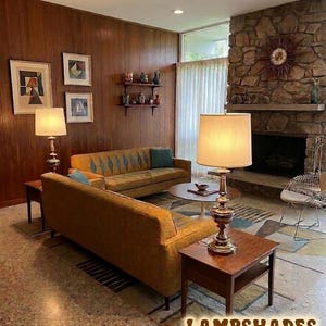 May include: A living room featuring two mustard yellow sofas, two wooden end tables with brass lamps, a round coffee table, and a stone fireplace. The walls are wood paneled with framed art. Text at the bottom reads: LAMPSHADES NOT INCLUDED.