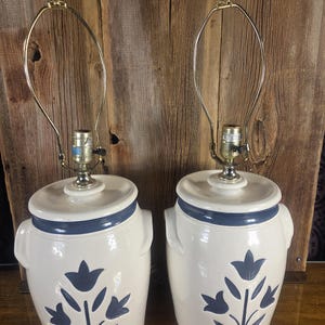 May include: Pair of ceramic lamps with a white base, blue floral design, and a blue rim. Each lamp has a metal neck and a silver finial. The lamps are designed to look like vintage crocks.
