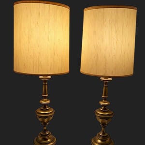 May include: A pair of brass table lamps with a tiered, pedestal-style base and a slender central column. Each lamp is topped with a cylindrical, textured off-white fabric shade. The text "Shades not included" is visible in the bottom right corner.