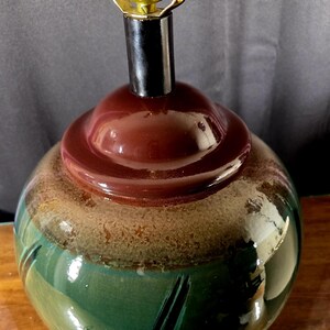 May include: A ceramic table lamp with a round, bulbous base. The base features a color gradient of green, tan, and brown. The lamp has a black neck and a brass-colored lamp holder.