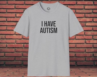 Può includere: T-shirt grigio chiaro con la scritta nera "I HAVE AUTISM". La maglietta ha maniche corte e girocollo. La maglietta è mostrata su uno sfondo di muro di mattoni.