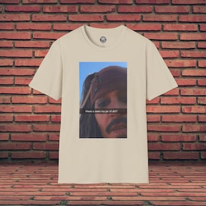 Op de afbeelding: Een beige t-shirt met een rechthoekige afbeelding van een persoon met een bruine bandana. De afbeelding bevat de tekst "Have u seen my jar of dirt?" in het wit. Het shirt heeft een ronde hals en korte mouwen.