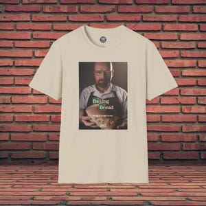 Könnte beinhalten: Ein beiges T-Shirt mit einer Grafik eines Mannes, der Brot hält, mit dem Text "Baking Bread". Das Shirt hat einen Rundhalsausschnitt und kurze Ärmel. Der Hintergrund ist eine Backsteinmauer.